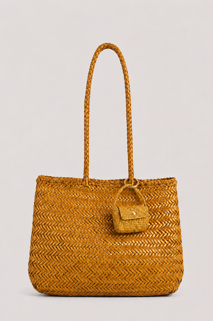 A mustard yellow woven straw tote bag with braided shoulder straps and a small matching woven front pouch, photographed against a solid light gray background.